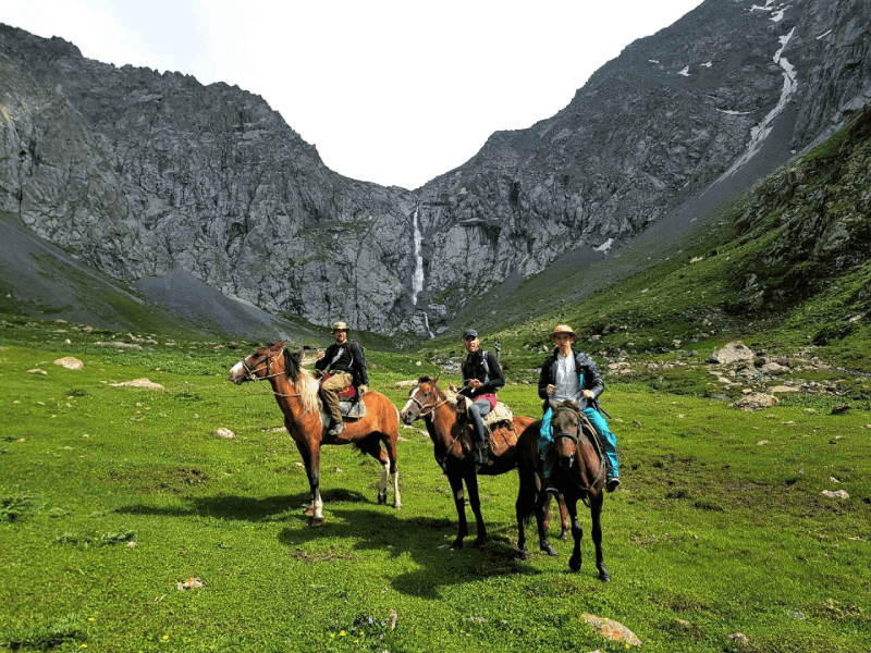 Three riders on horseback in a green valley below a waterfall cascading down grey cliffs